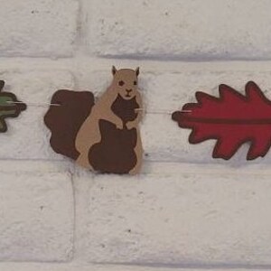 May include: A garland with alternating brown squirrels and green and red leaf shapes. The garland is made of paper and is strung together with string.