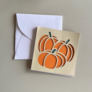 以下が含まれることがあります： 切り抜かれたカボチャのデザインが特徴の正方形のグリーティングカード。緑色の茎が付いた3つのオレンジ色のカボチャが描かれています。カードはベージュ色で、オレンジ色の背景があり、白い封筒が添えられています。秋や感謝祭に最適です。