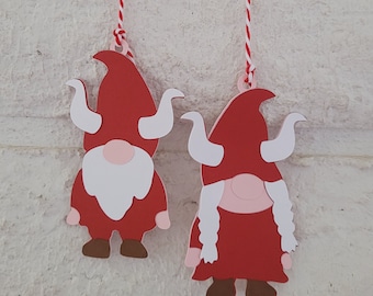 May include: Two red felt gnome ornaments with white accents hanging from red and white twine. The gnome on the left has a white beard and a red hat. The gnome on the right has white hair and a red hat.