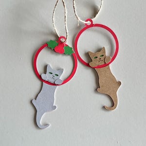 May include: Two cat-shaped Christmas ornaments. One is gray, the other is tan. Both cats are hanging from red circles. The gray cat ornament has green eyes and is decorated with a red holly berry and green leaves. The tan cat has green eyes.