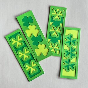 Puede incluir: Cuatro marcapáginas de papel verde y amarillo con diseños de tréboles. Cada marcapáginas tiene forma rectangular con borde verde e interior amarillo. Los diseños de tréboles son en varios tonos de verde.