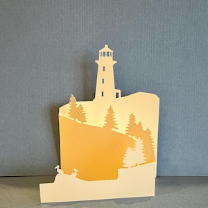 Puede incluir: Una obra de arte en papel en capas que presenta un faro en un acantilado, con árboles y pájaros. La obra de arte utiliza tonos crema, amarillo y naranja, creando una escena cálida. El fondo es azul apagado.