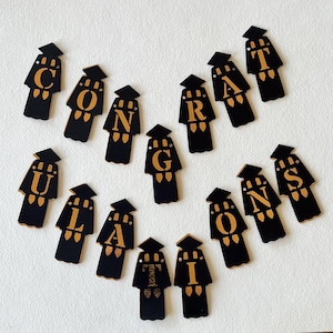 Puede incluir: Recortes de gorras de graduación negros y dorados que deletrean "CONGRATULATIONS". Cada recorte tiene la forma de una gorra y una toga de graduación, con letras doradas. Los recortes están dispuestos sobre una superficie blanca.