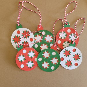 Könnte beinhalten: Sechs rote und grüne Weihnachtsbaumschmuck-Geschenk-Etiketten mit weißen Schneeflocken- und Sternmotiven. Jede Etikette hat eine weiße Schnur.
