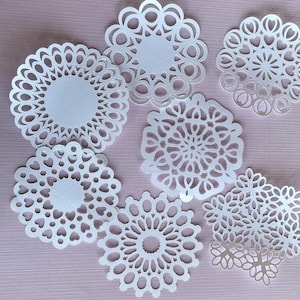 May include: A collection of white paper doilies in various intricate floral and geometric designs. The doilies are arranged on a light pink surface, showcasing their delicate, openwork patterns. The designs include heart shapes and circular patterns.