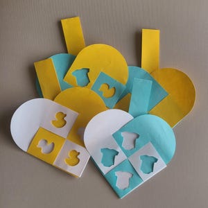 Op de afbeelding: Drie papieren hartvormige labels met uitgesneden ontwerpen. Een label is geel met een eend en een rompertje, een is wit met een eend en een rompertje, en een is blauw met een rompertje.