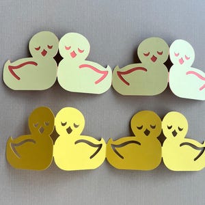 May include: Eight paper cut-out ducklings in various shades of yellow and green. Each duckling has a curved wing detail and closed eyes. The ducklings are arranged in pairs, with red accents on the wings and beaks.