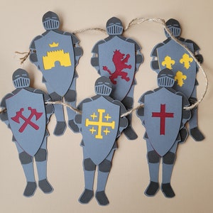 Peut inclure: Une guirlande de chevaliers en papier gris, chacun avec un bouclier orné d'emblèmes héraldiques. On y trouve un château jaune, un lion rouge, des fleurs de lys jaunes, des haches rouges, une croix jaune et une croix rouge. Les chevaliers sont reliés par de la ficelle.