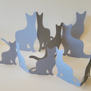 Puede incluir: Un grupo de recortes de papel de gatos en varias poses, dispuestos en un círculo. Los gatos son todos grises, algunos son de un tono de gris más claro que otros.