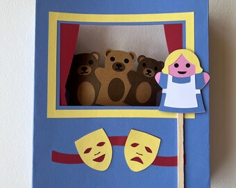 May include: A handmade paper theater scene with three brown teddy bear puppets on a stage with red curtains. A blonde girl puppet is on a stick. The theater is blue with yellow and red accents, and includes happy and sad face masks.