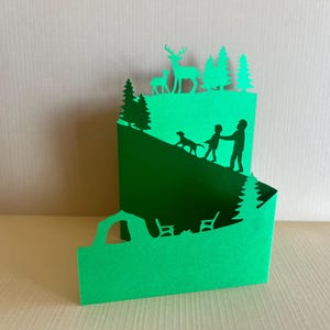 Puede incluir: Una pieza de arte en papel verde en capas que representa una escena de ladera. Siluetas de una persona, un niño y un perro ascienden una colina, con ciervos y árboles en la cima. La capa inferior muestra un vehículo y bancos.