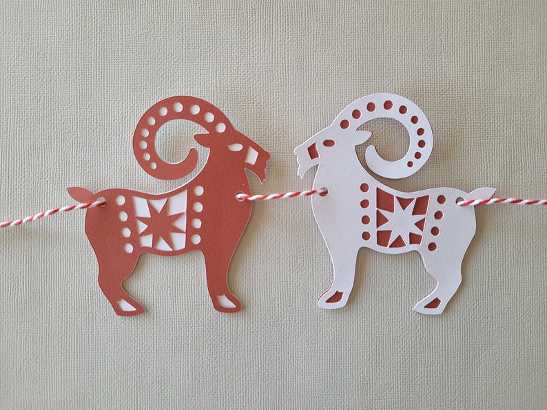 May include: A red and white paper garland with two cut-out goats connected by a red and white string. The goats have a star and geometric pattern.
