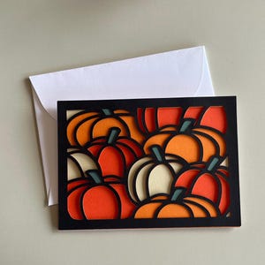 Può includere: Una carta nera con un design a strati di zucche nei toni dell'arancione, rosso e crema. Le zucche hanno steli verdi. Una busta bianca è dietro la carta, adatta per l'autunno o il Ringraziamento.