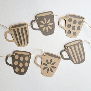 Puede incluir: Una pancarta de tazas de café de papel marrón con diferentes patrones, incluyendo rayas, lunares y flores. Las tazas están enhebradas juntas con un cordel.