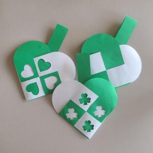 Op de afbeelding: Drie groene en witte papieren hartvormige enveloppen met uitgesneden klavertjevier-ontwerpen. De enveloppen zijn perfect voor St. Patrick's Day.