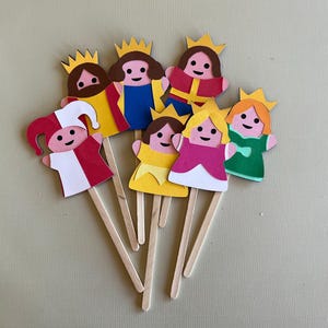 Puede incluir: Una colección de coloridas muñecas de papel sobre palitos de madera. Las muñecas presentan varios atuendos, incluyendo coronas, vestidos y un gorro de bufón. La paleta de colores incluye rojo, amarillo, azul, rosa y verde. Probablemente para manualidades o juegos infantiles.
