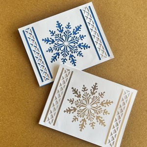 May include: Two white cards with snowflake cutouts. One card features a blue snowflake and blue border accents, while the other has a gold snowflake and gold border accents. The cards are rectangular and likely for a winter holiday.