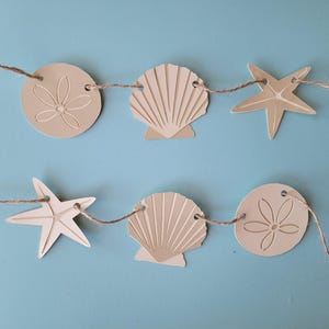May include: A beige and brown paper garland with alternating seashells, starfish, and sand dollar shapes. The garland is strung together with twine.