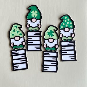 May include: Four St. Patrick's Day gnome cutouts. Each gnome has a green hat with a shamrock or polka dot design, a white beard, and a pink nose. The gnomes are standing on stacks of books with black and white pages.