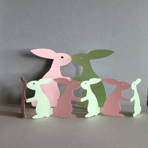 May include: A collection of colorful paper cut-out bunnies in shades of pink, green, and brown. The bunnies are arranged in a group, with some holding hands. The background is a solid gray.