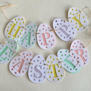 Puede incluir: Una guirnalda de Pascua con recortes en forma de huevo que deletrean "HAPPY EASTER". Cada huevo es blanco con lunares de colores y letras en amarillo, verde, rosa y morado. La guirnalda está ensartada con hilo.