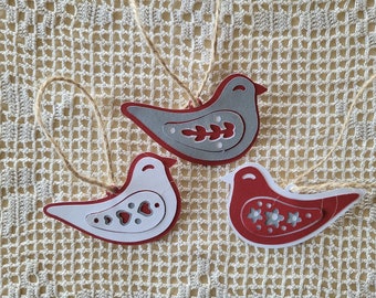 May include: Three paper bird ornaments with a rustic twine hanger. The birds are layered with white paper on the inside and red or gray paper on the outside. Each bird has a different design cut out of the center.