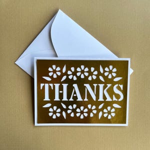 Könnte beinhalten: Eine goldfarbene "THANKS"-Grußkarte mit floralem Design und einem weißen Umschlag. Die Karte hat einen weißen Rand und ist vor einem goldenen Hintergrund platziert. Die Karte ist eine Dankeskarte.