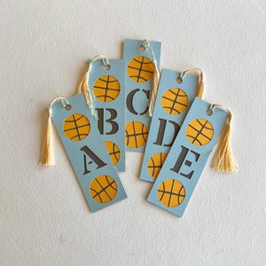 May include: Five light blue rectangular bookmarks, each featuring a dark gray letter from A to E and yellow basketball graphics. Each bookmark has a hole at the top with a cream-colored tassel attached, creating a set of sports-themed markers.