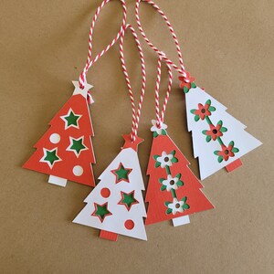 Puede incluir: Cuatro etiquetas de regalo de árbol de Navidad rojas y blancas con diseños de estrellas verdes y flores. Cada etiqueta tiene una cuerda blanca.