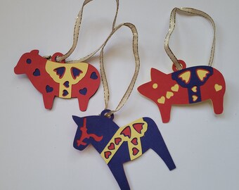 May include: Three Christmas ornaments, a red sheep, a red pig, and a blue horse, each with yellow and red hearts. The ornaments are made of paper and have gold string for hanging.