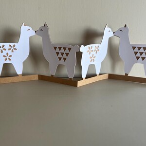 May include: Four white and gray llama figurines kissing, standing on a brown base. Two llamas have flower cutouts, and two have triangle cutouts. The figurines are a decorative item.