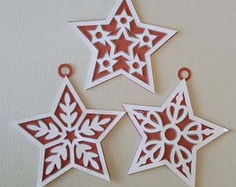 May include: Three white paper star ornaments with intricate cut-out designs. The stars are layered with a red-brown paper backing.