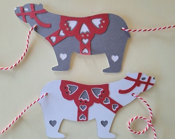 May include: Two paper bear ornaments, one gray and one white, are connected by red and white twine. Each bear has a red bow and heart cutouts. The bears are on a light yellow background.
