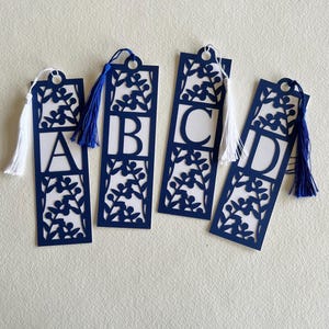 Puede incluir: Cuatro marcadores de libros de papel azul marino con recortes florales blancos y las letras A, B, C y D. Cada marcador tiene una borla, con borlas blancas y azules alternas. Los marcadores son rectangulares con un agujero circular en la parte superior.