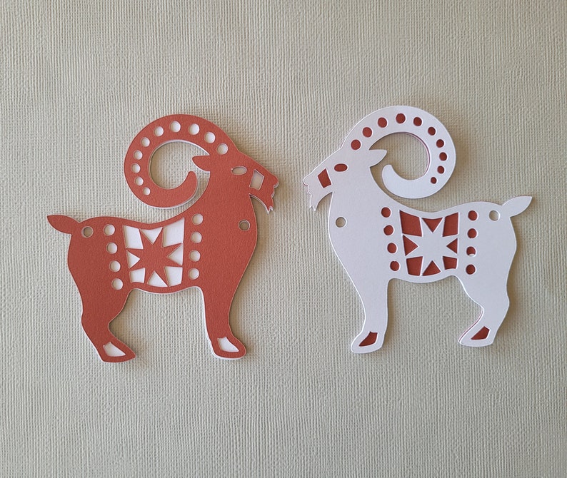 May include: Two paper cutouts of goats, one red and one white, with a star design. The goats are facing opposite directions.
