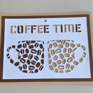 Pode incluir: Um design de estêncil com tema de café com as palavras "COFFEE TIME" acima de duas canecas de café cheias de grãos de café. O design está em um fundo branco com uma borda marrom.