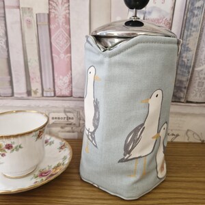 May include: A duck egg blue tea cosy featuring a seagull design, alongside a floral teacup and saucer. The cosy has a stainless steel top and a black knob. The background shows a bookshelf wallpaper.
