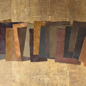 May include: Assortment of 14 leather swatches in various shades of brown, tan, and dark brown. The leather pieces are arranged in a row on a wooden surface.