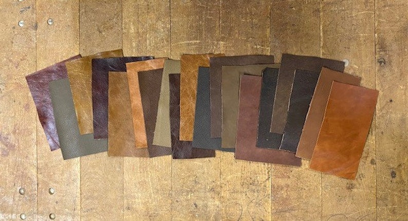 May include: Assortment of 14 leather swatches in various shades of brown, tan, and dark brown. The leather pieces are arranged in a row on a wooden surface.