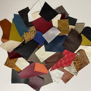 May include: Assortment of leather scraps in various colors and textures, including brown, red, blue, black, white, and beige. The leather pieces are arranged in a pile on a white surface.