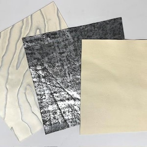 May include: Three square sheets of material with distinct textures. One sheet features a light cream color with wavy, embossed lines. The second sheet is dark gray with a mottled, distressed pattern. The third sheet is a solid, light beige color.