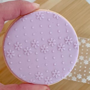 May include: A round cookie with a light purple frosting decorated with small flowers and dots. The cookie is held by a hand. A clear, square stamp with a flower pattern is next to the cookie.
