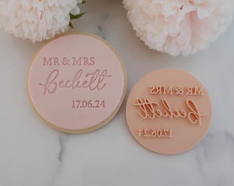 Pode incluir: Um biscoito rosa claro com as palavras "MR & MRS Beckett 17.06.24" impressas, ao lado de um carimbo rosa claro com o mesmo texto invertido. O biscoito é redondo e tem uma borda marrom clara. O fundo inclui flores brancas e rosas.
