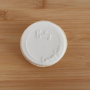 K&ouml;nnte beinhalten: Ein runder Keks mit wei&szlig;em Zuckerguss. Der Zuckerguss tr&auml;gt die Worte "Baby" in Schreibschrift oben und "Coming Soon" unten. Der Keks liegt auf einer Holzoberfl&auml;che.