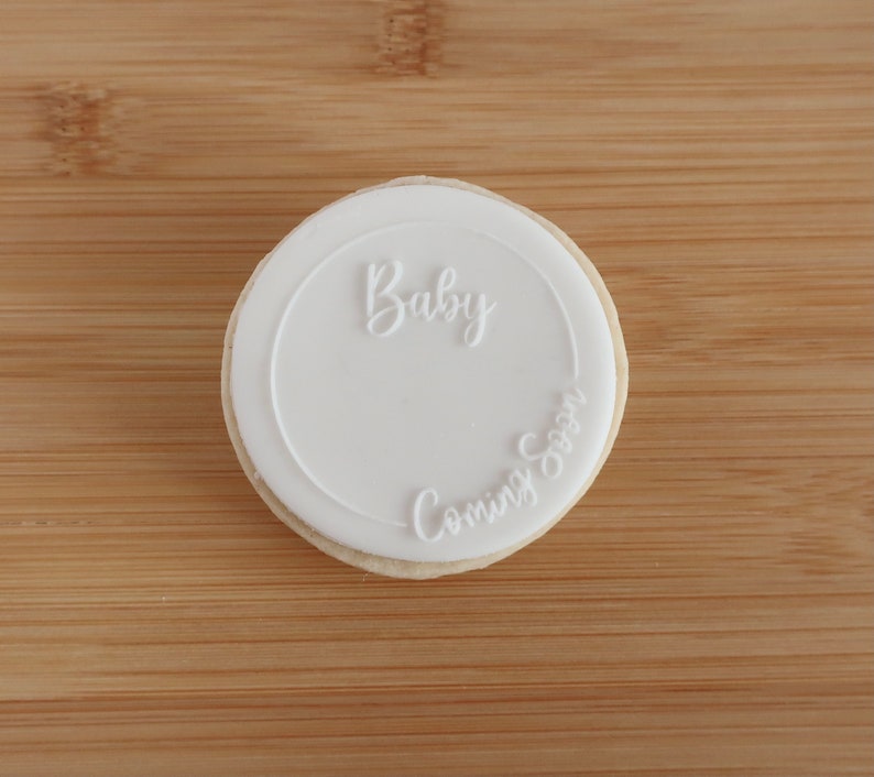 K&ouml;nnte beinhalten: Ein runder Keks mit wei&szlig;em Zuckerguss. Der Zuckerguss tr&auml;gt die Worte "Baby" in Schreibschrift oben und "Coming Soon" unten. Der Keks liegt auf einer Holzoberfl&auml;che.
