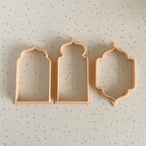 May include: Three light peach-colored cookie cutters in different arched window shapes. The cutters are made of plastic and are arranged side by side on a speckled surface. The shapes include a rounded arch, a pointed arch, and a quatrefoil.