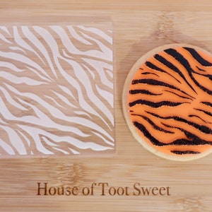 May include: A clear plastic stencil featuring a white zebra stripe design, alongside a round biscuit decorated with orange icing and black stripes, mimicking tiger fur. The words "House of Toot Sweet" are visible.