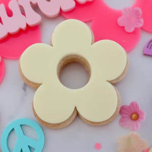 May include: A flower-shaped biscuit with a white icing centre and a light brown biscuit base. The biscuit is surrounded by pink and light blue decorative elements, including a peace sign and flower shapes. The overall theme is a retro, floral design.
