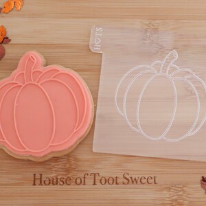 May include: A pumpkin-shaped cookie cutter and a decorated cookie. The cookie is light orange with a pumpkin design. The clear plastic cutter has a pumpkin outline. The text "House of Toot Sweet" is visible. Decorative fall leaves are scattered around.