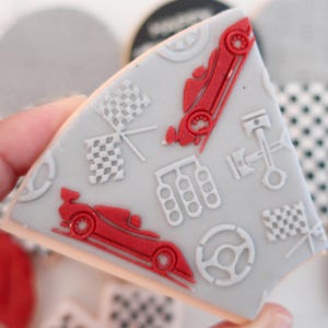 May include: A grey, triangular biscuit decorated with red race cars, chequered flags, and a steering wheel. Other biscuits with similar designs are visible in the background. The biscuit is held by a hand.
